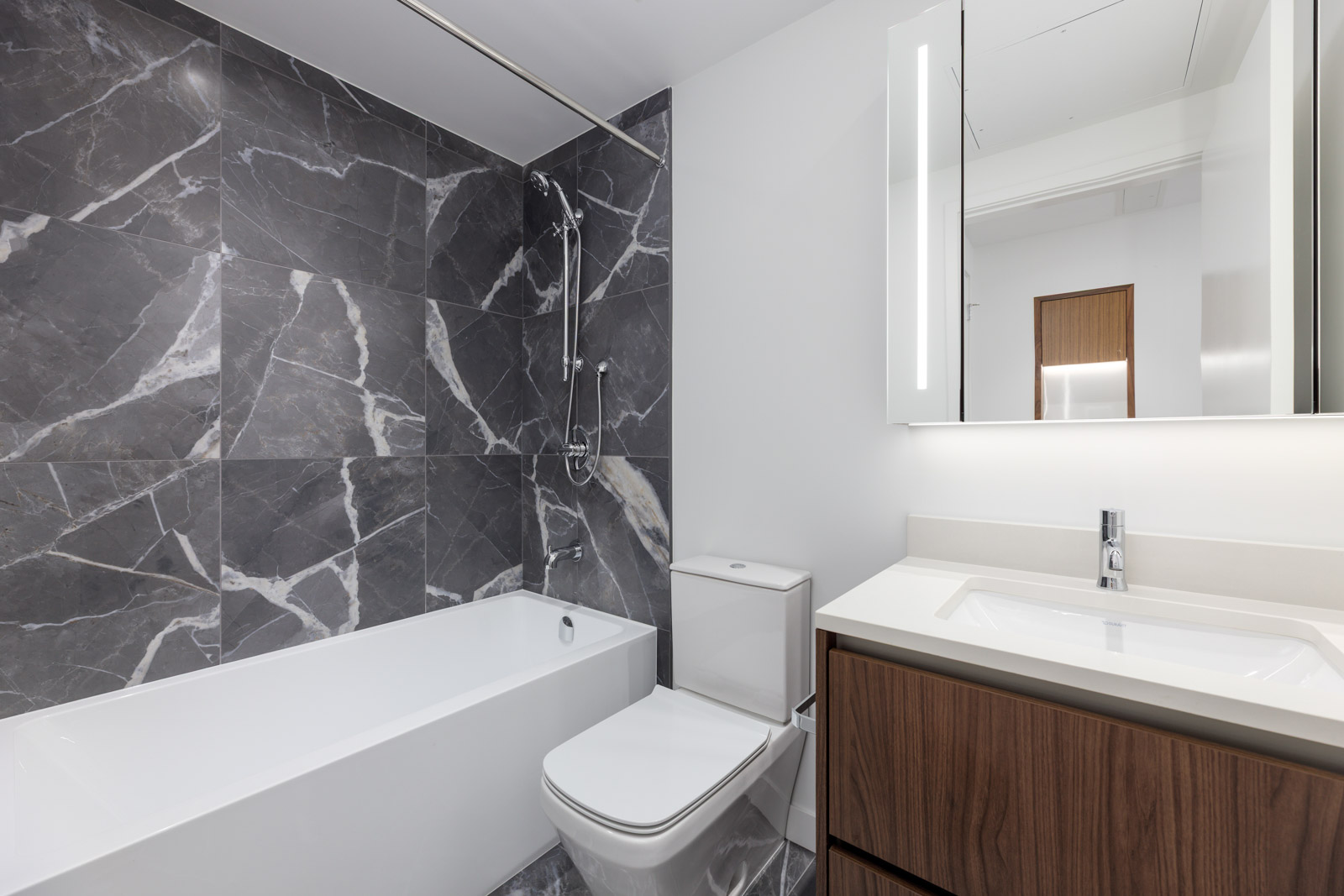 Modern bathroom with gray marble tile walls, a white bathtub with shower, a toilet, a wooden vanity with sink, and a mirrored cabinet above the sink.