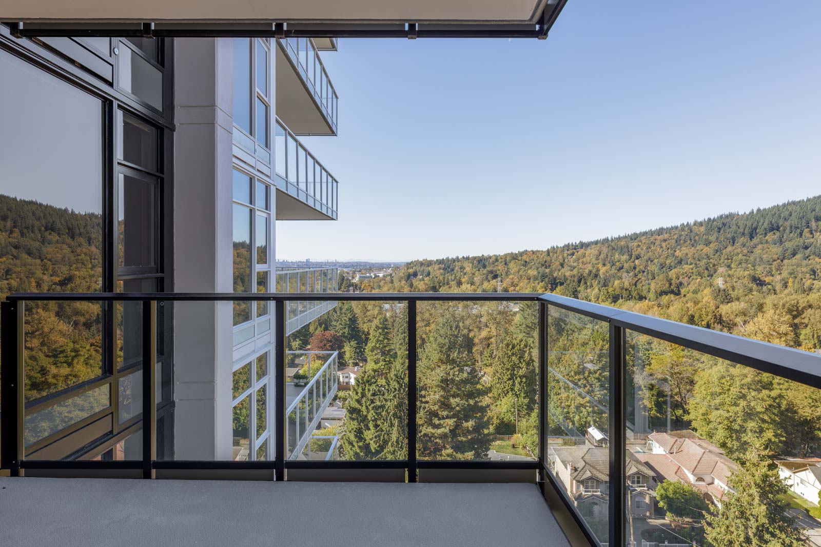 Glass balcony of a modern high-rise building overlooking a forested hillside and residential neighborhood under a clear sky.