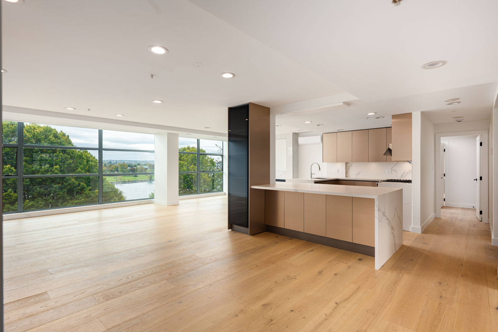 Modern open-plan kitchen and living area with light wood flooring, large windows, and views of greenery and water outside.
