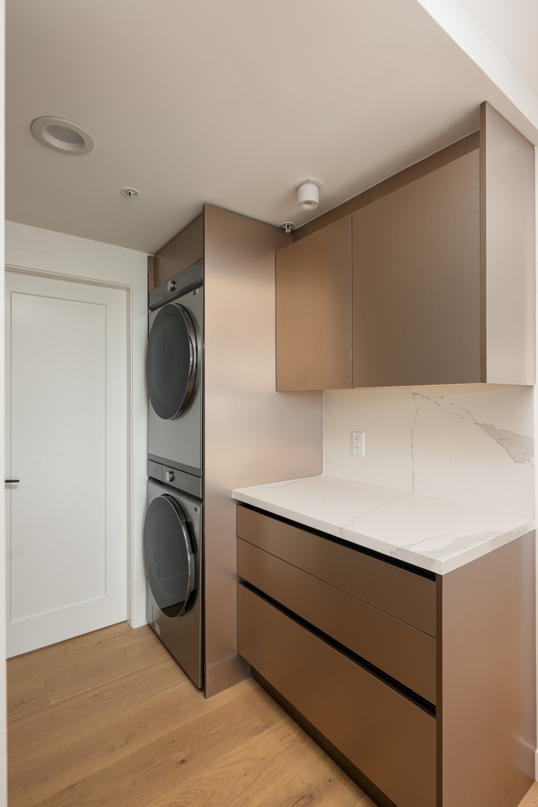 Modern laundry room with a stacked washer and dryer, brown cabinets, white marble countertop, and light wood flooring.