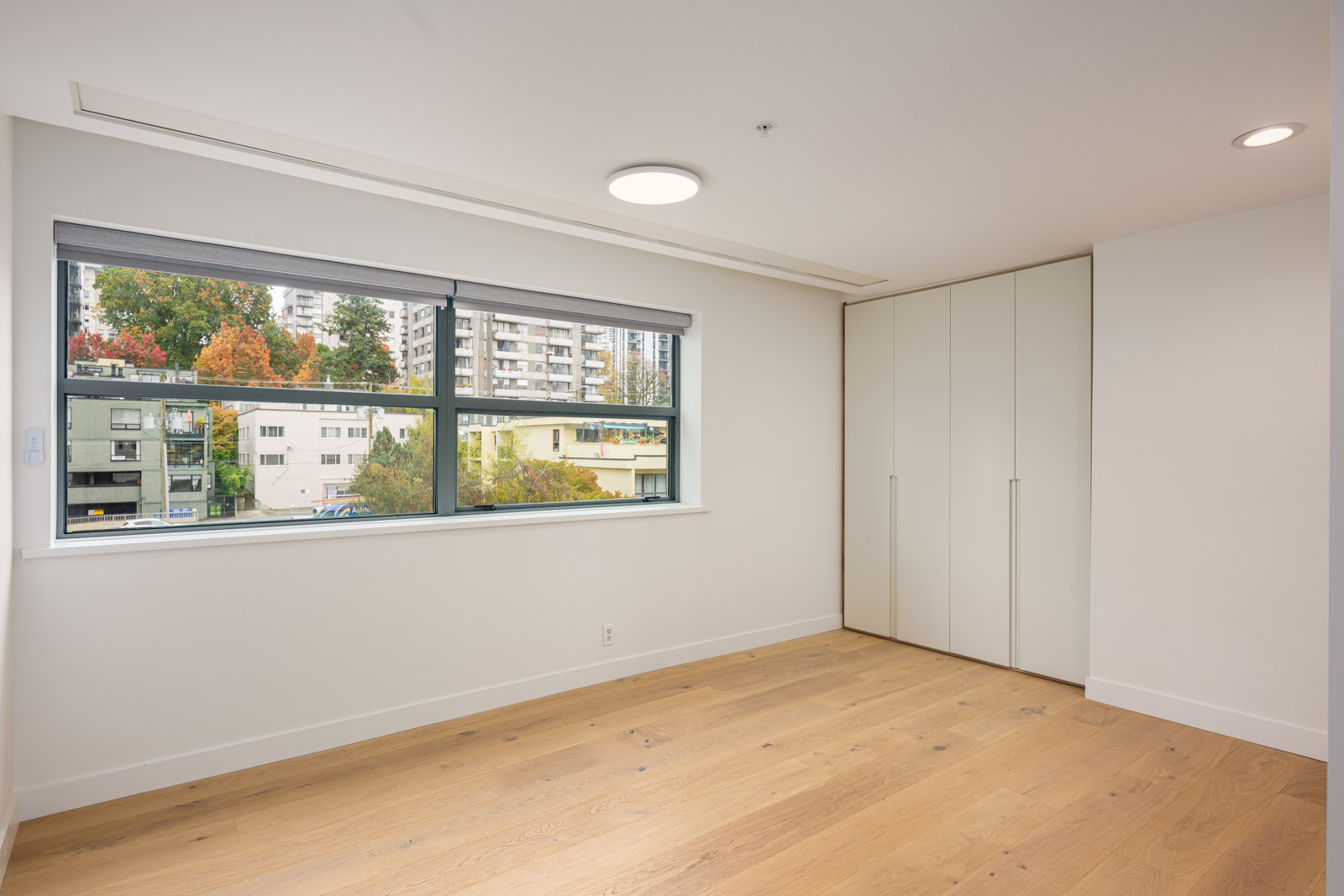 Empty room with light wood flooring, large window showing buildings and trees outside, white walls, and a closed built-in closet.