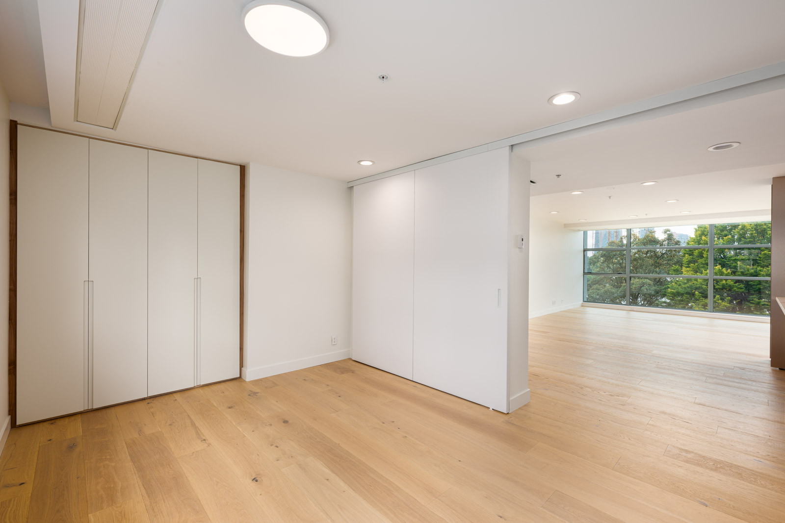 Modern, empty room with light wooden floors, white walls, a closed closet, recessed lighting, and large windows overlooking greenery in the adjacent space.
