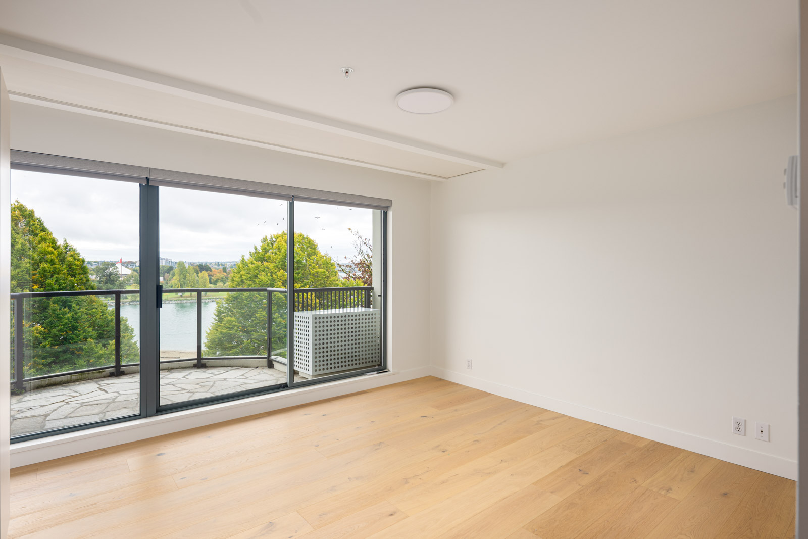 Empty room with light wood floors, white walls, and large sliding glass doors opening to a balcony with a lake and trees visible outside.