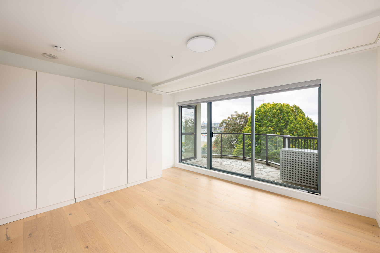 A bright, empty room with light wood flooring, built-in white cabinets, and large sliding glass doors leading to a balcony with trees visible outside.