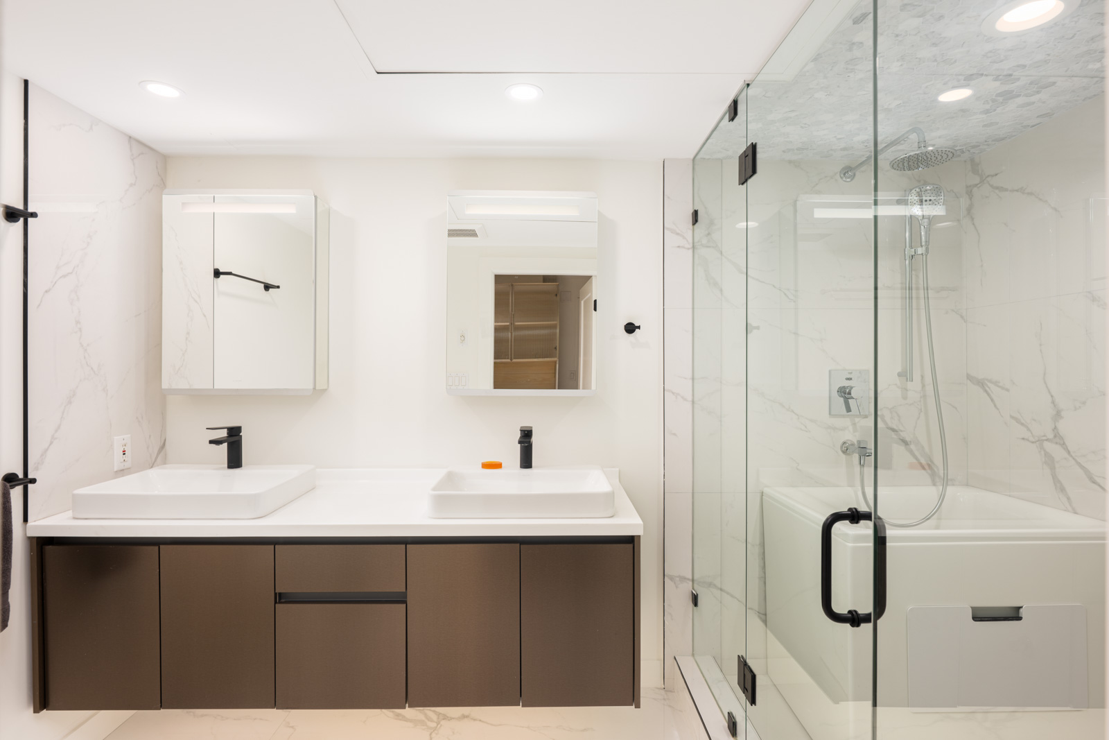 Modern bathroom with double sinks, marble walls, brown cabinetry, large mirrors, and a glass-enclosed shower-tub combination.