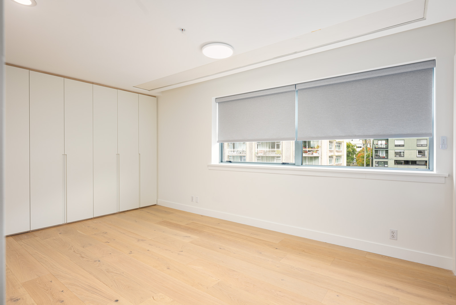 Empty, unfurnished room with light wood flooring, white walls, large built-in closets, and a window with grey blinds partially open to reveal an exterior view.