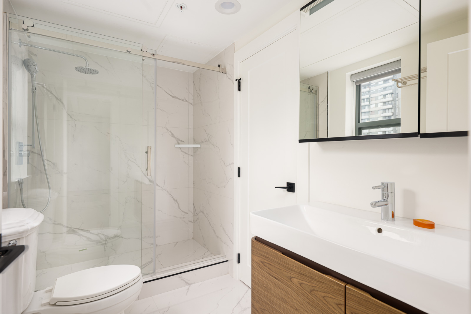 Modern bathroom with a glass-enclosed shower, wall-mounted toilet, wide sink with wood vanity, mirrored cabinet, and a window providing natural light.