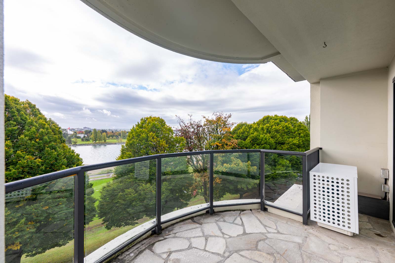 Curved balcony with glass railing overlooks trees, grass, and a body of water under a cloudy sky. A white utility box is placed near the wall.