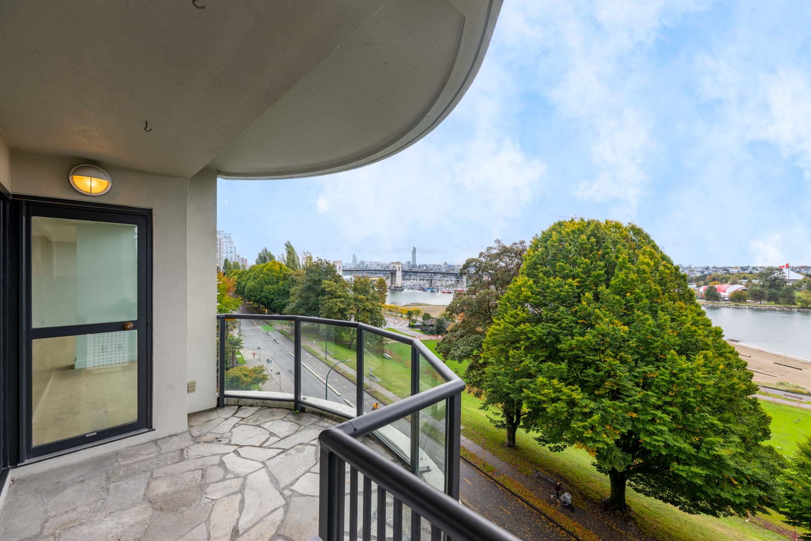 A balcony with stone flooring overlooks a park, trees, a walkway, and a body of water with a bridge visible in the background under a partly cloudy sky.