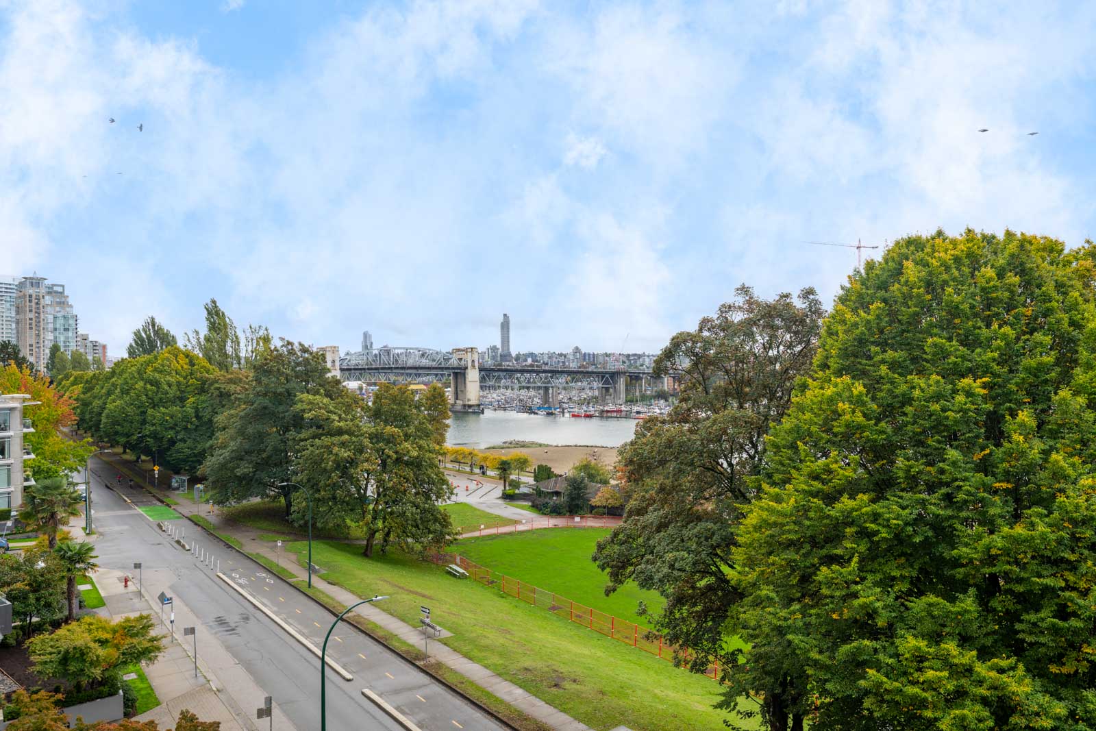 A city street runs alongside a park with green trees, leading to a bridge over a river under a partly cloudy sky.