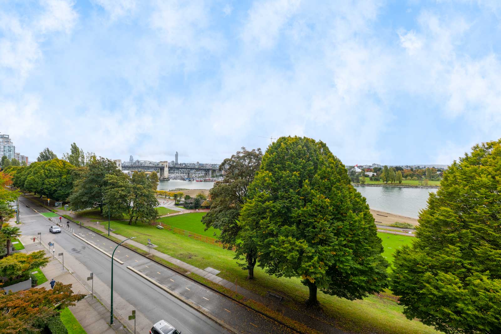 A city street runs alongside a green park with large trees, next to a river with a bridge in the background under a partly cloudy sky.