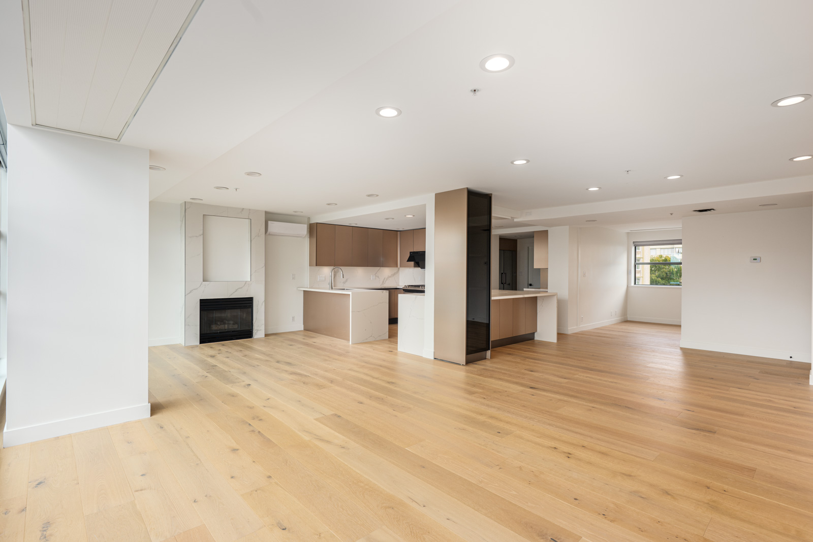 Spacious modern open-plan living area with light wood flooring, recessed lighting, a fireplace, and a kitchen with light brown cabinets. Large windows provide natural light.