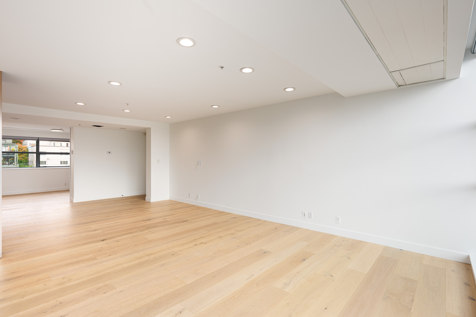 Empty modern room with light wood flooring, white walls, recessed ceiling lights, and large windows letting in natural light.