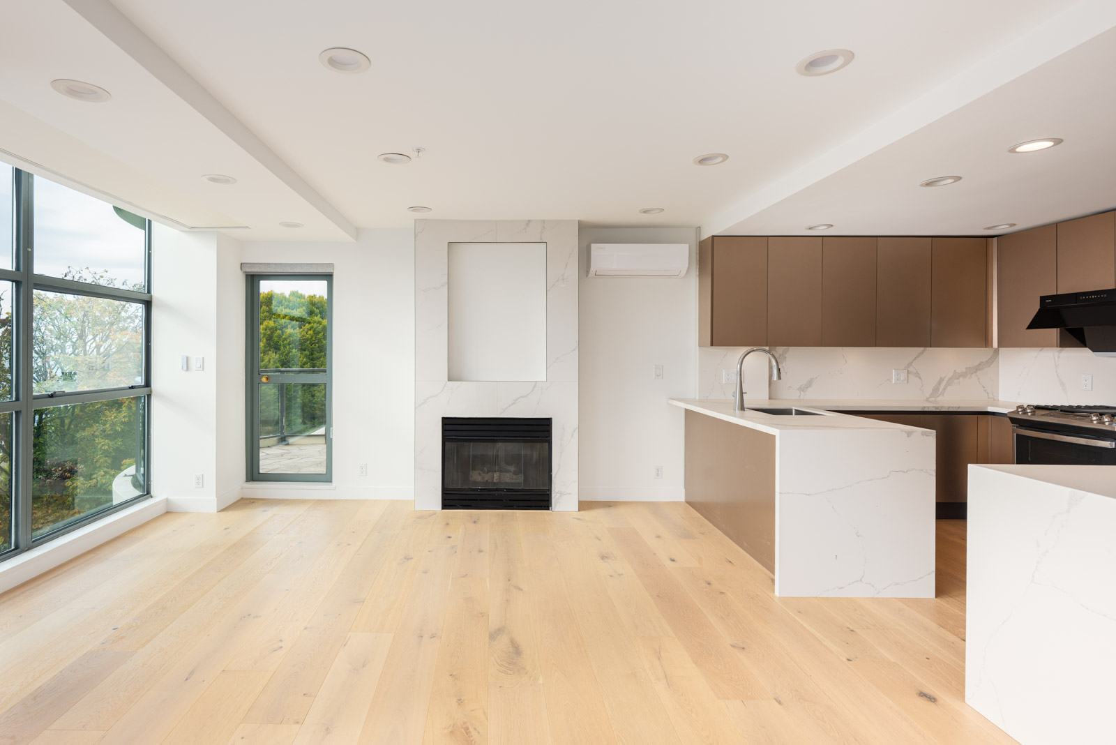 Modern, unfurnished apartment with large windows, light wood flooring, a fireplace, marble accents, and an open kitchen with brown cabinets.