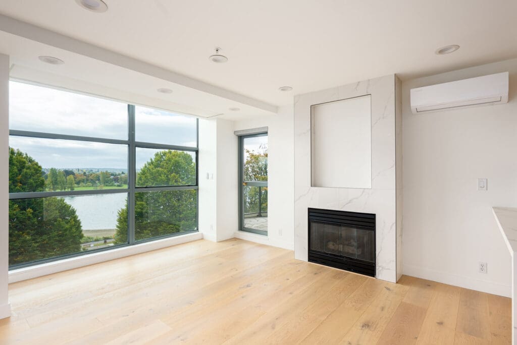 A bright, empty living room with large windows overlooking trees and water, light wood floors, a modern fireplace, and white walls.
