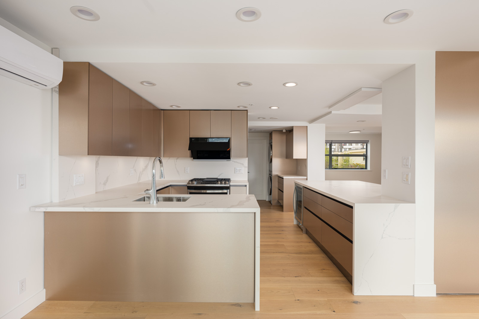 Modern kitchen with brown cabinets, white marble countertops, built-in appliances, and light wood flooring, featuring recessed ceiling lights and large windows in the background.