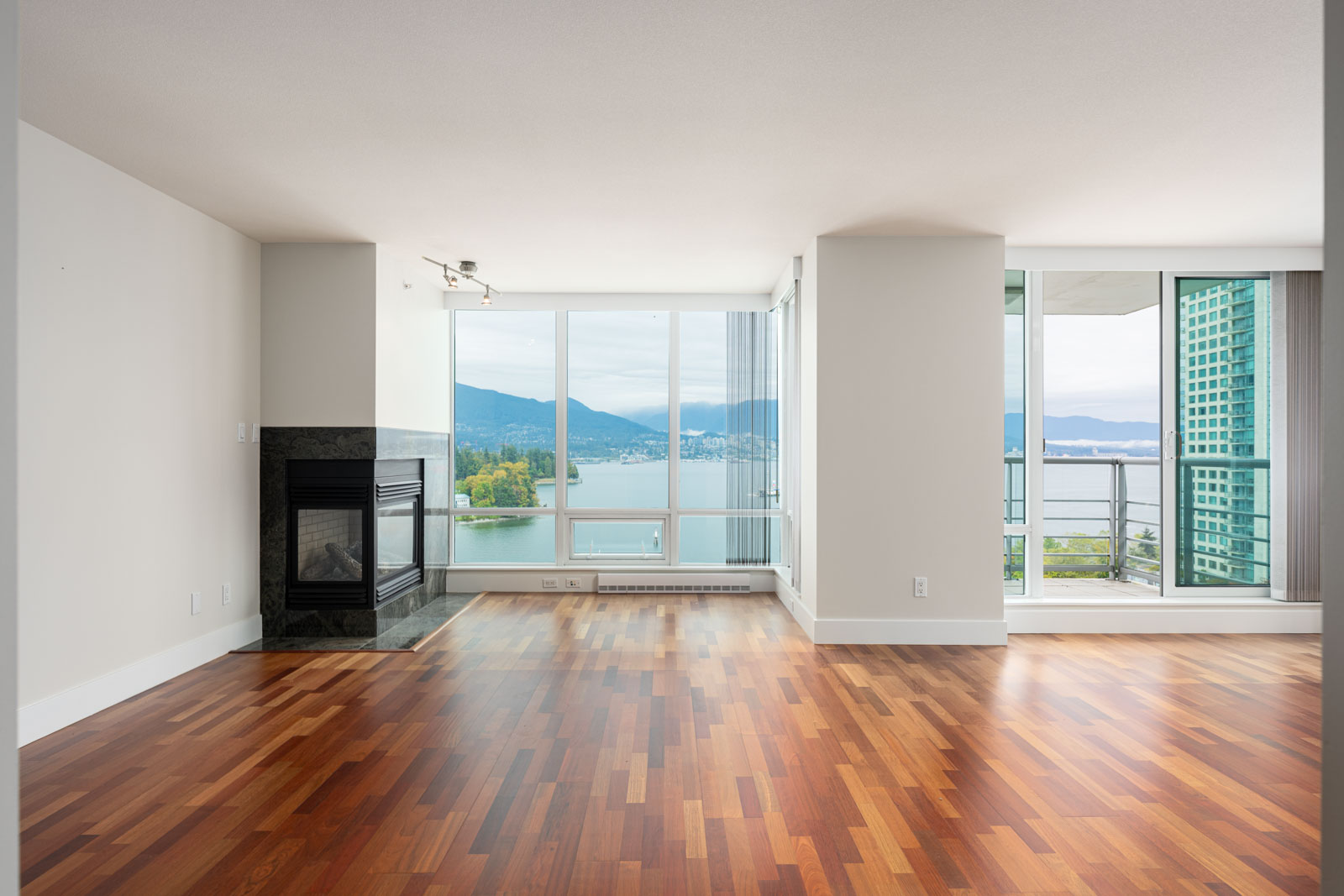 Unfurnished apartment living room with hardwood floors, large windows, and a fireplace, overlooking a balcony with a view of water and mountains.