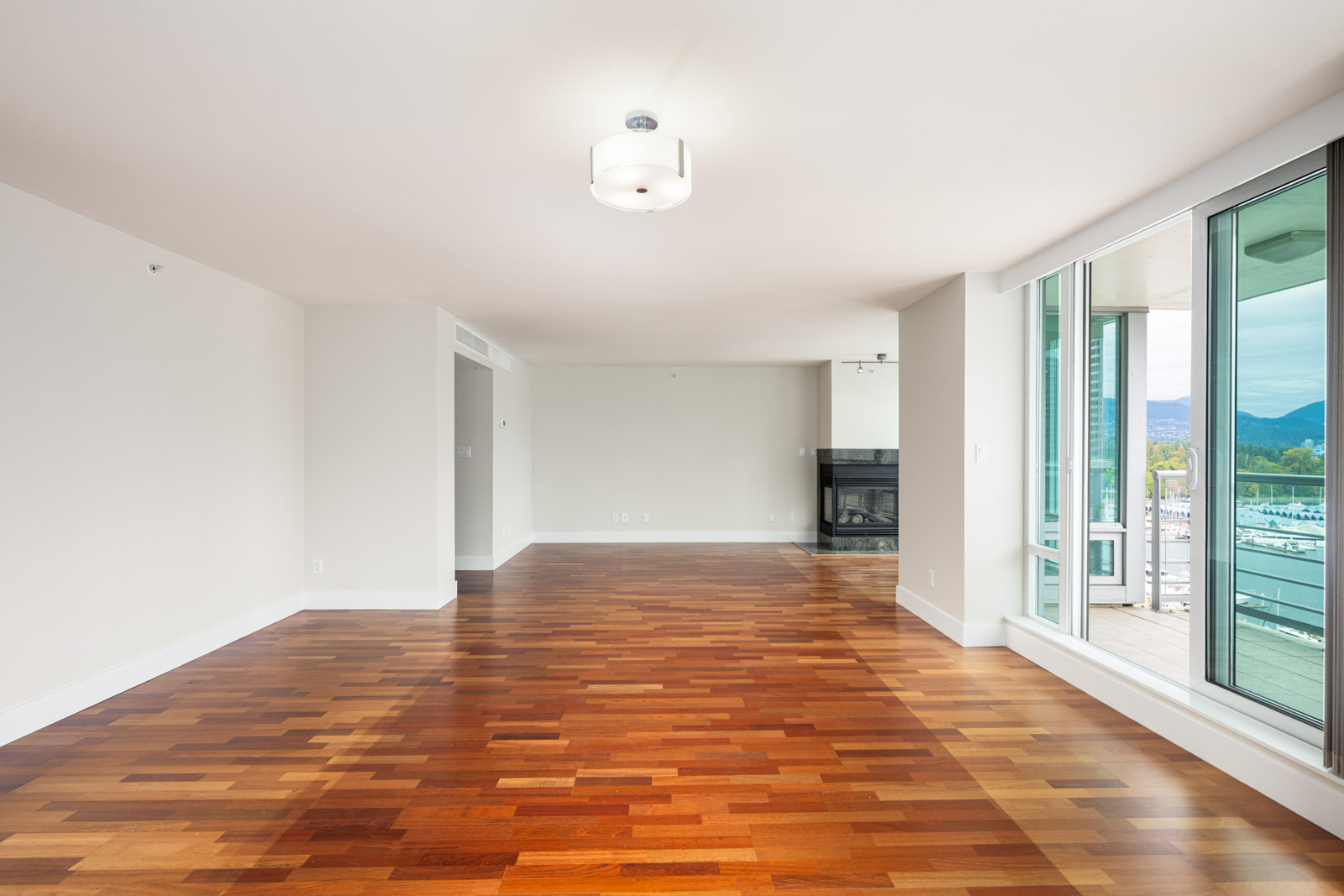 Spacious empty living room with hardwood floors, white walls, ceiling light fixture, large windows, and glass door opening to a balcony with an outdoor view.