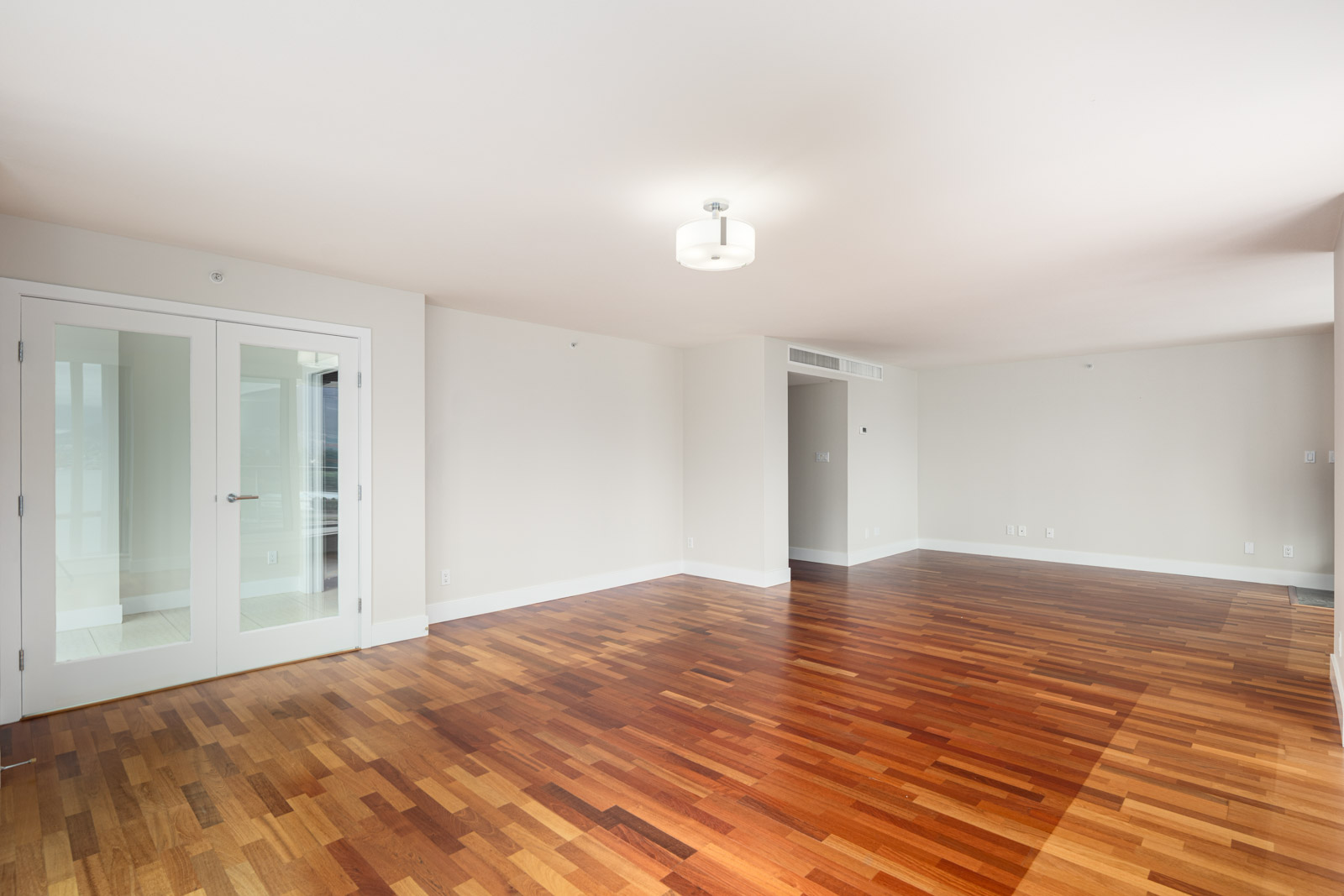 Spacious, empty room with hardwood floors, white walls, a ceiling light fixture, and double glass doors leading to another area.