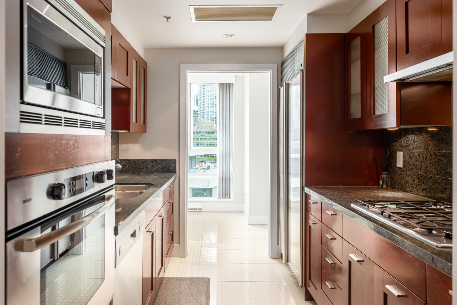 Modern galley kitchen with stainless steel appliances, dark wood cabinets, and a window at the far end letting in natural light, overlooking city buildings.