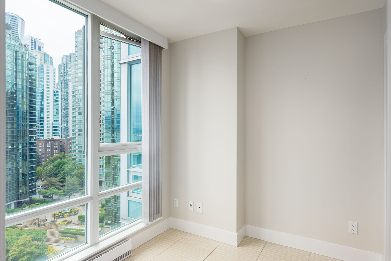 A corner of a modern apartment with large windows, beige walls, and a view of city buildings and greenery outside.