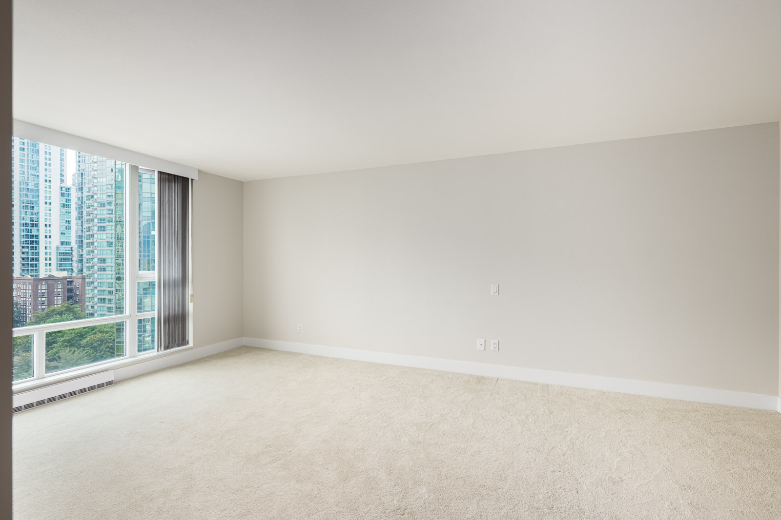 Empty carpeted room with large window showing city buildings and greenery; white walls and minimal electrical outlets visible.