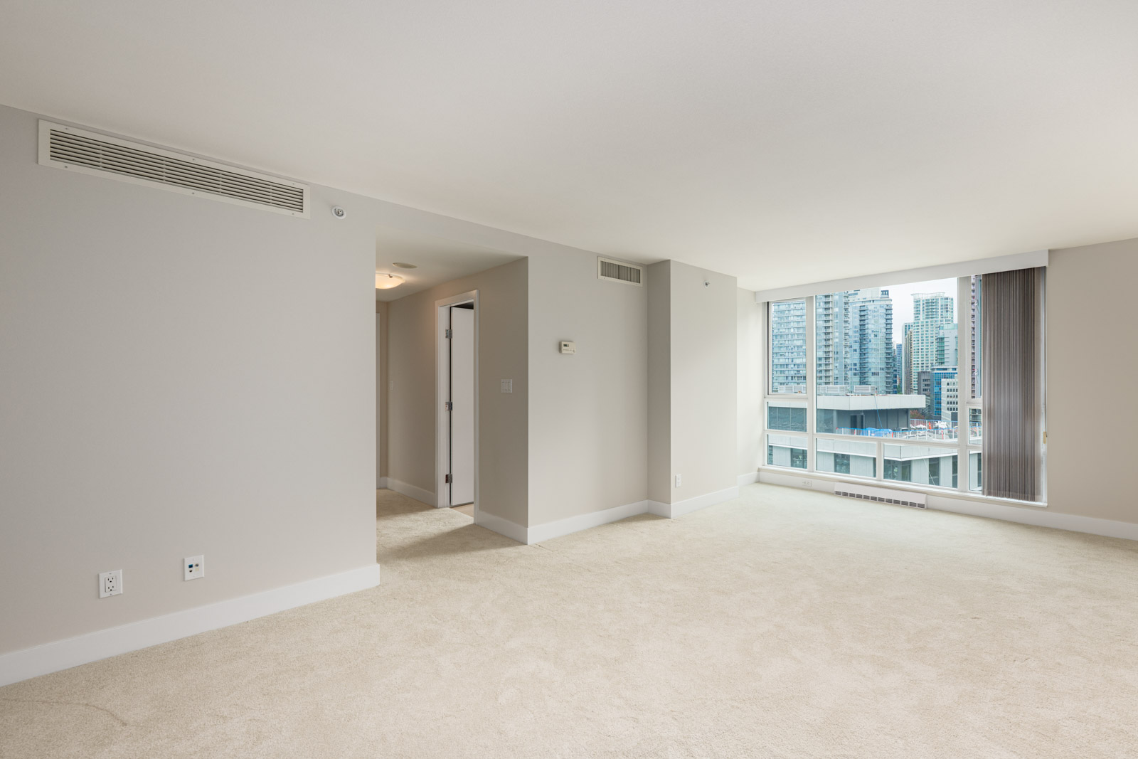 Empty, carpeted apartment room with beige walls, large window offering a city view, and a hallway leading to other areas.