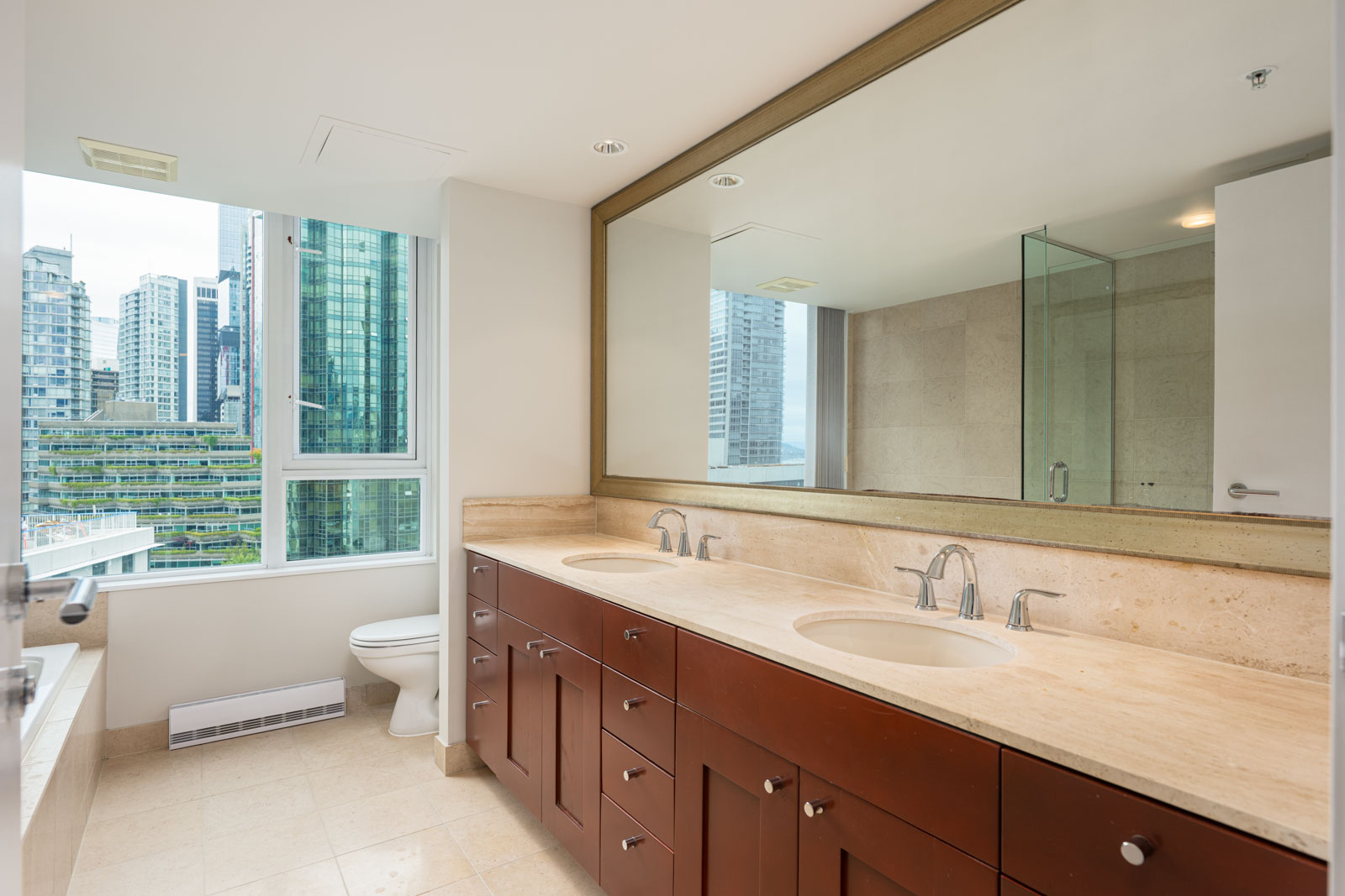 Modern bathroom with a large mirror, double sink vanity, glass shower, and a window offering a city view with tall buildings.