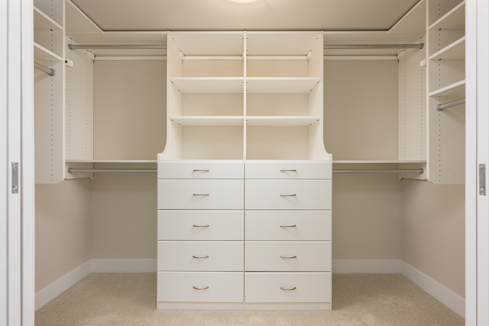 Empty walk-in closet with white shelves, drawers, and hanging rods on beige walls and carpeted floor.