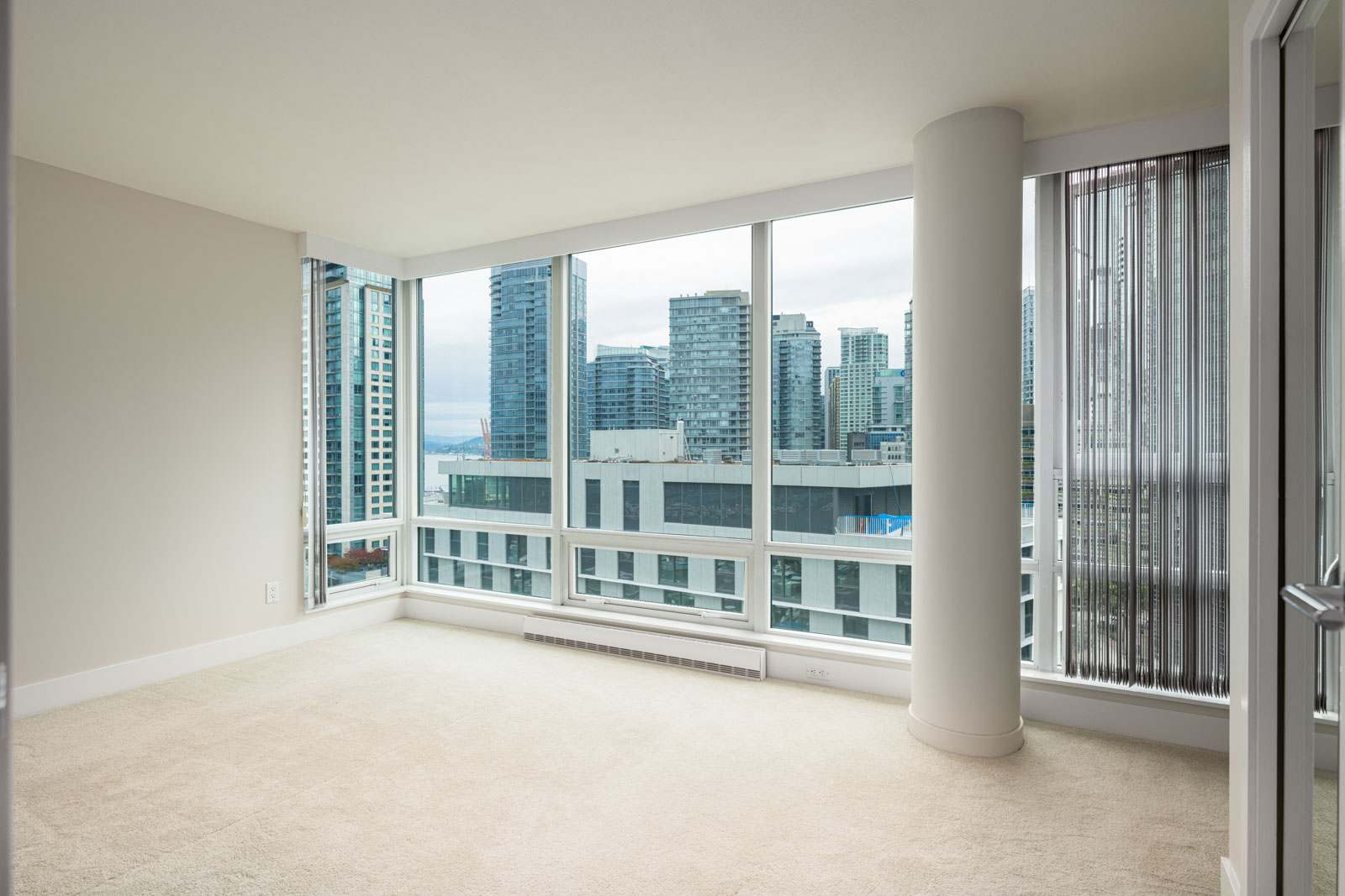 Empty corner room with floor-to-ceiling windows, beige carpet, white walls, and a view of city buildings and a cloudy sky outside.