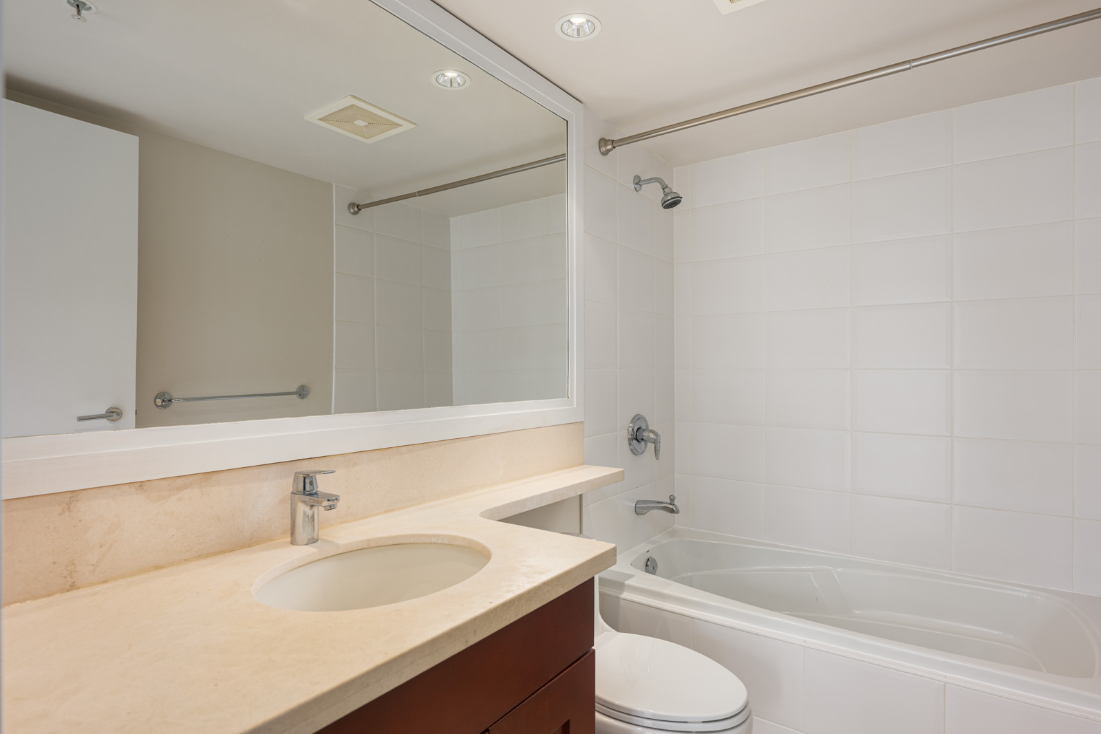 Modern bathroom with a large mirror, beige countertop with sink, wooden vanity, white bathtub with shower, and white tiled walls.