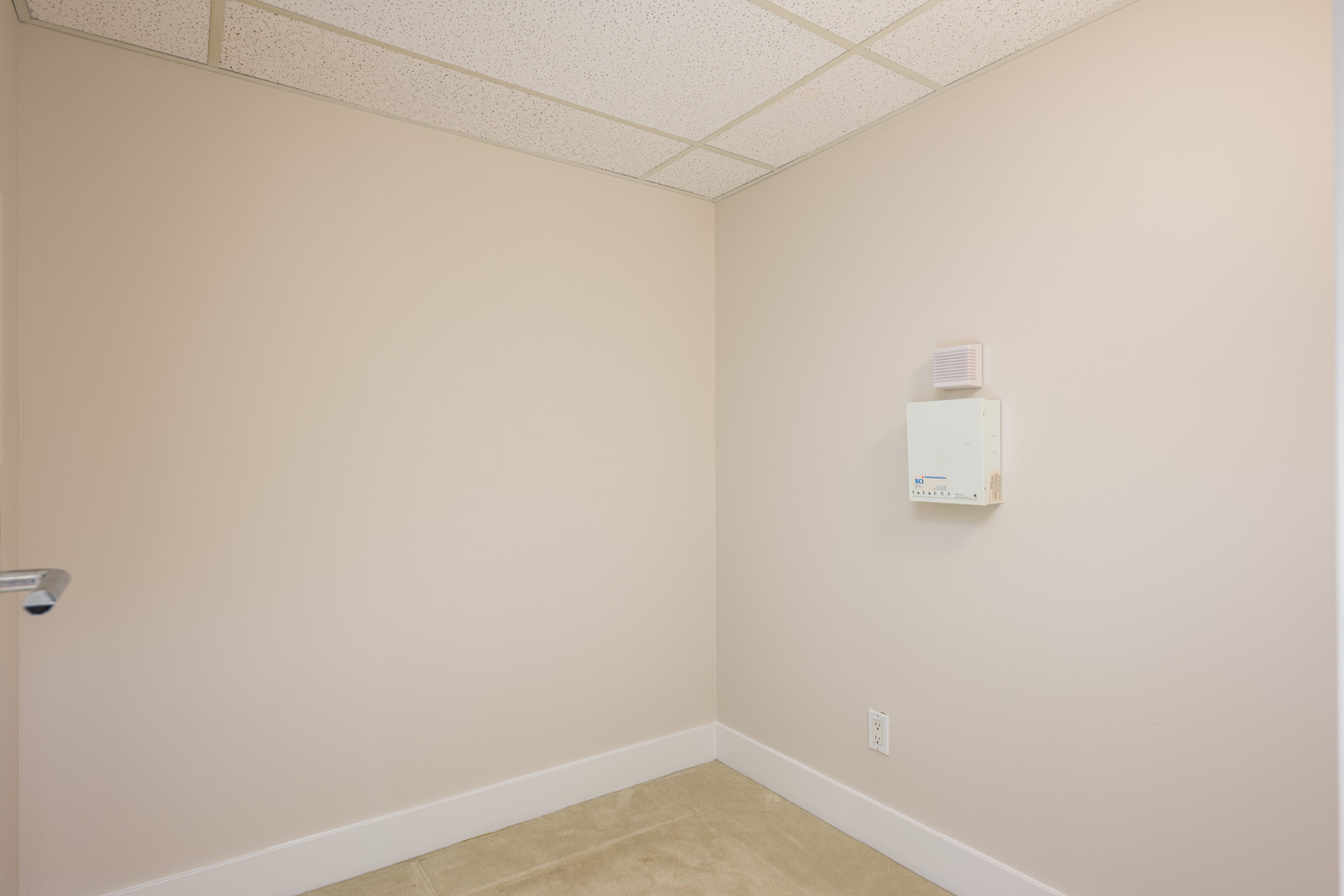 An empty beige room with a white baseboard, tiled ceiling, and a wall-mounted electrical box above an outlet.