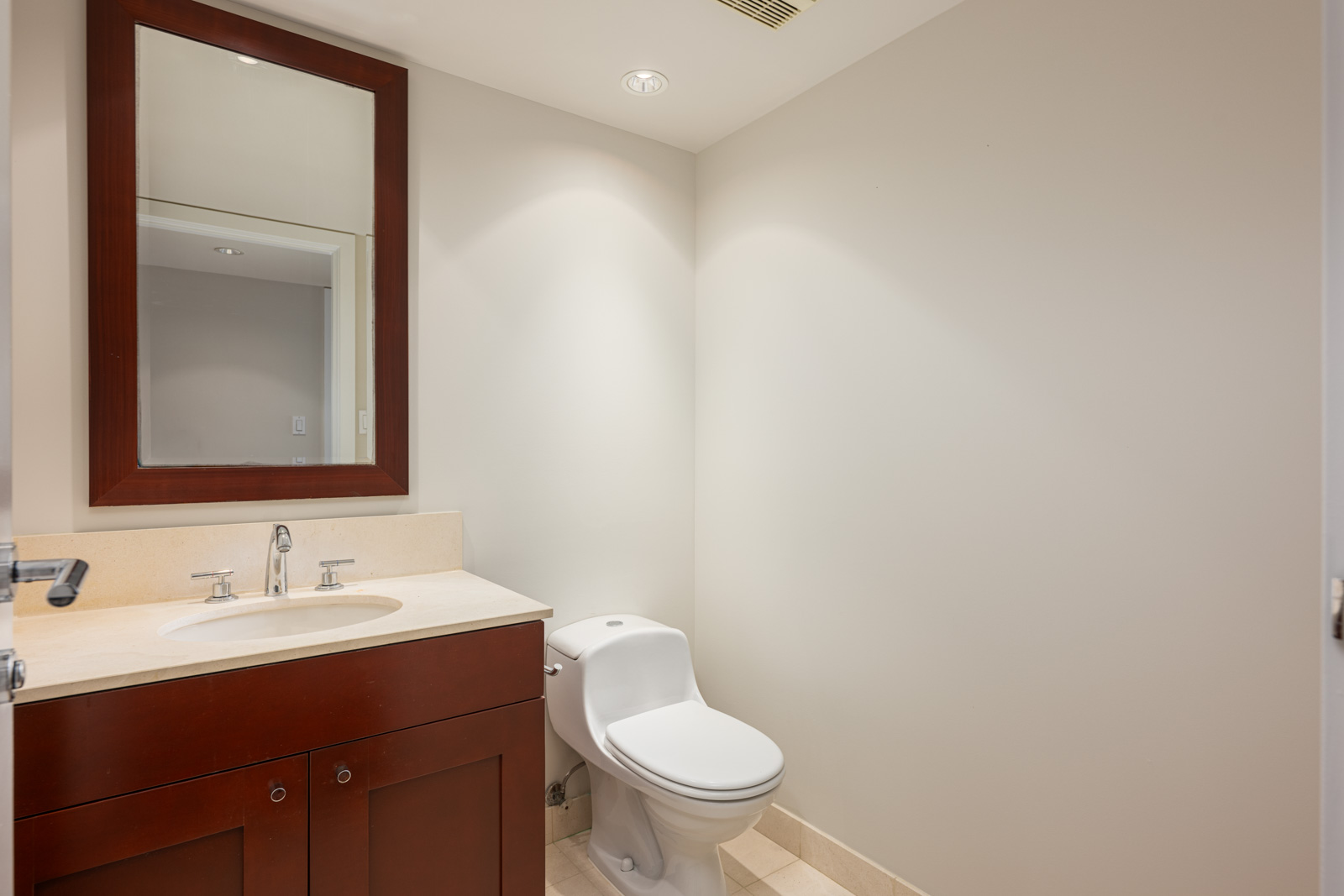 A bathroom with a sink and wooden vanity, a wall-mounted mirror, a toilet, and beige tiled floor under recessed ceiling lights.