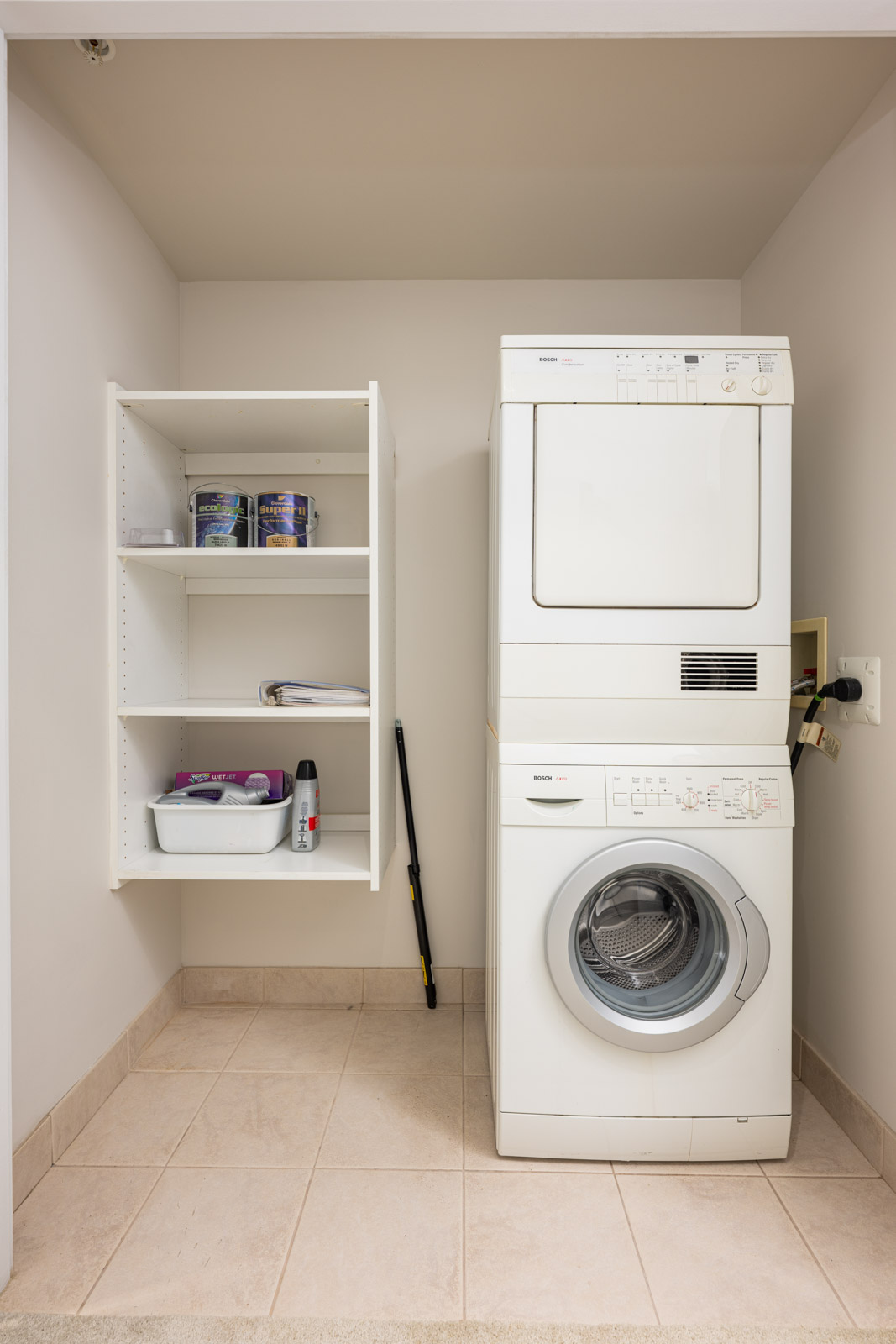 A white stacked washer and dryer next to a wall-mounted shelf holding paint cans, a tray, and cleaning supplies in a small laundry area with tiled flooring.