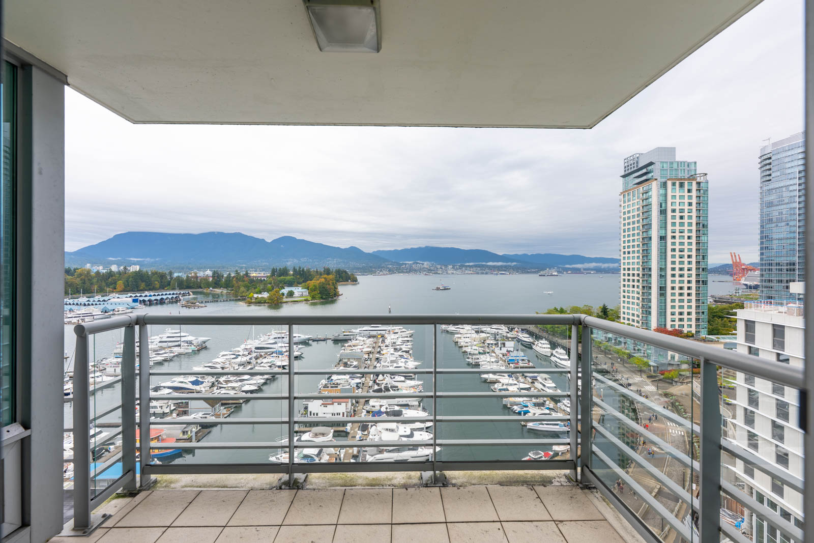 View from a balcony overlooking a marina with docked boats, tall buildings on the right, water, and mountains in the background under a cloudy sky.