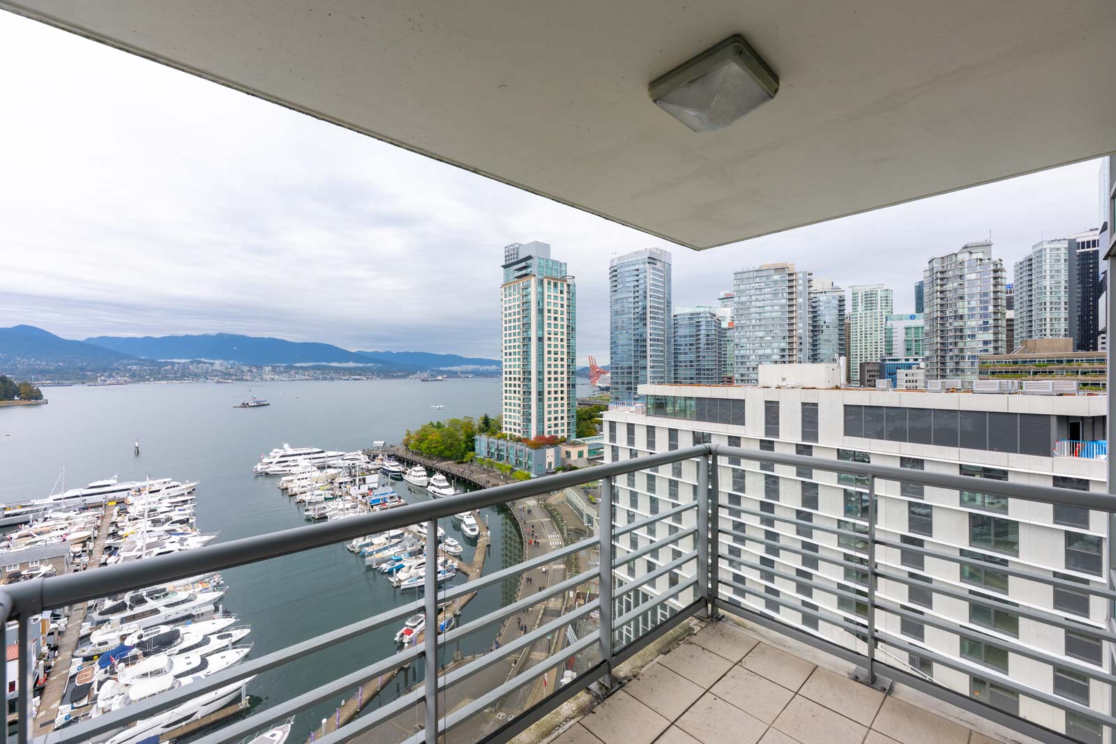 View from a high-rise balcony overlooking a marina, waterfront, and modern city buildings with mountains in the background under a cloudy sky.