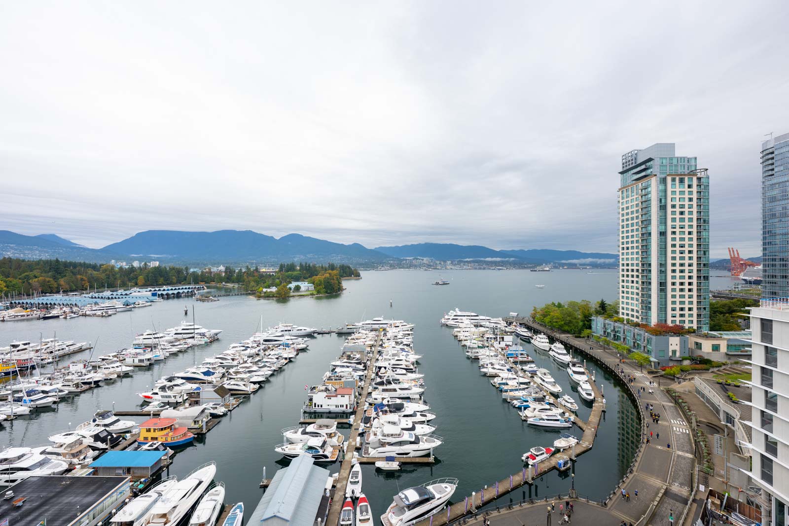 A marina filled with docked boats and yachts, bordered by tall modern buildings and a walkway, with mountains and cloudy sky in the background.