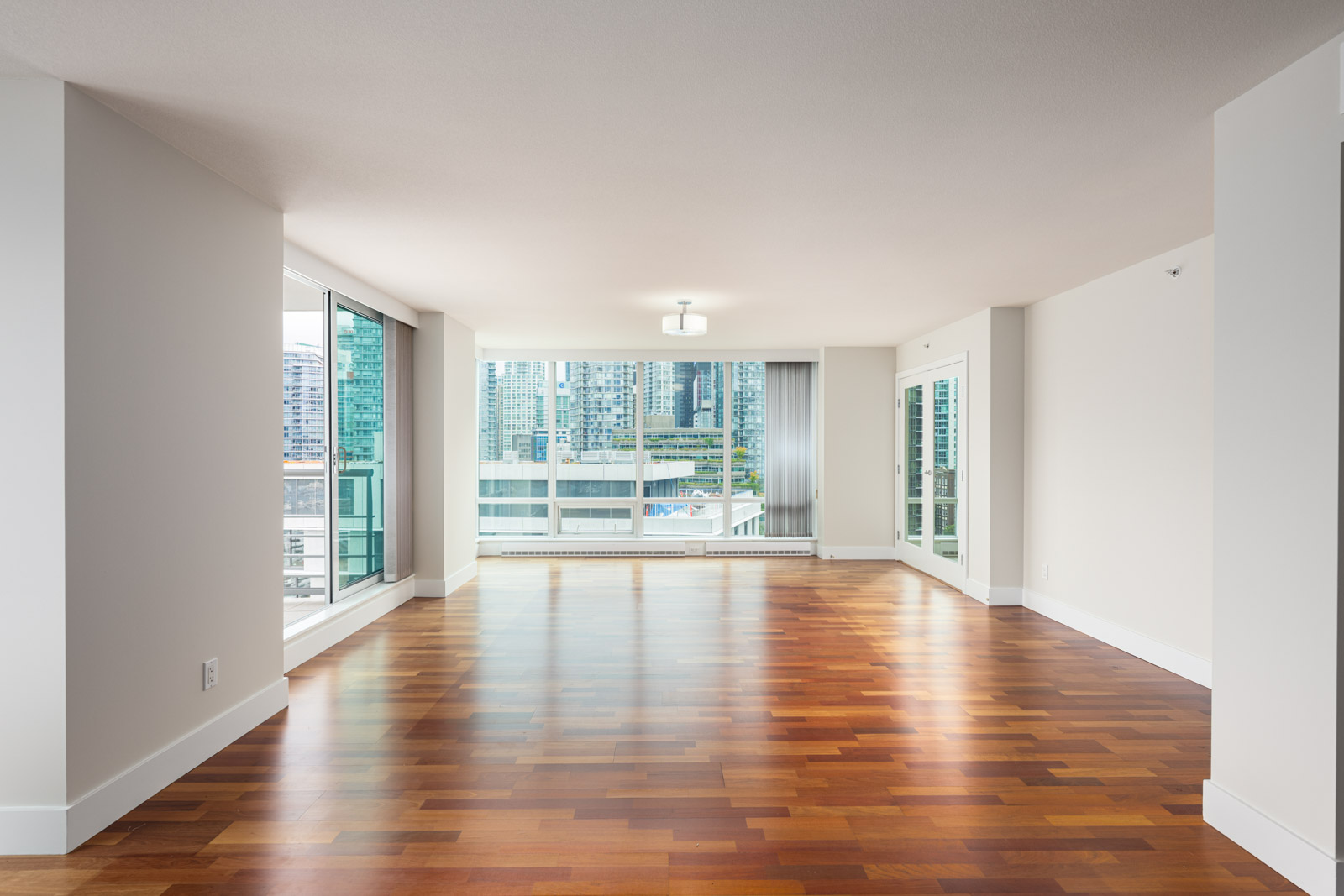 Spacious empty living room with hardwood floors, large windows, and a view of city buildings outside.