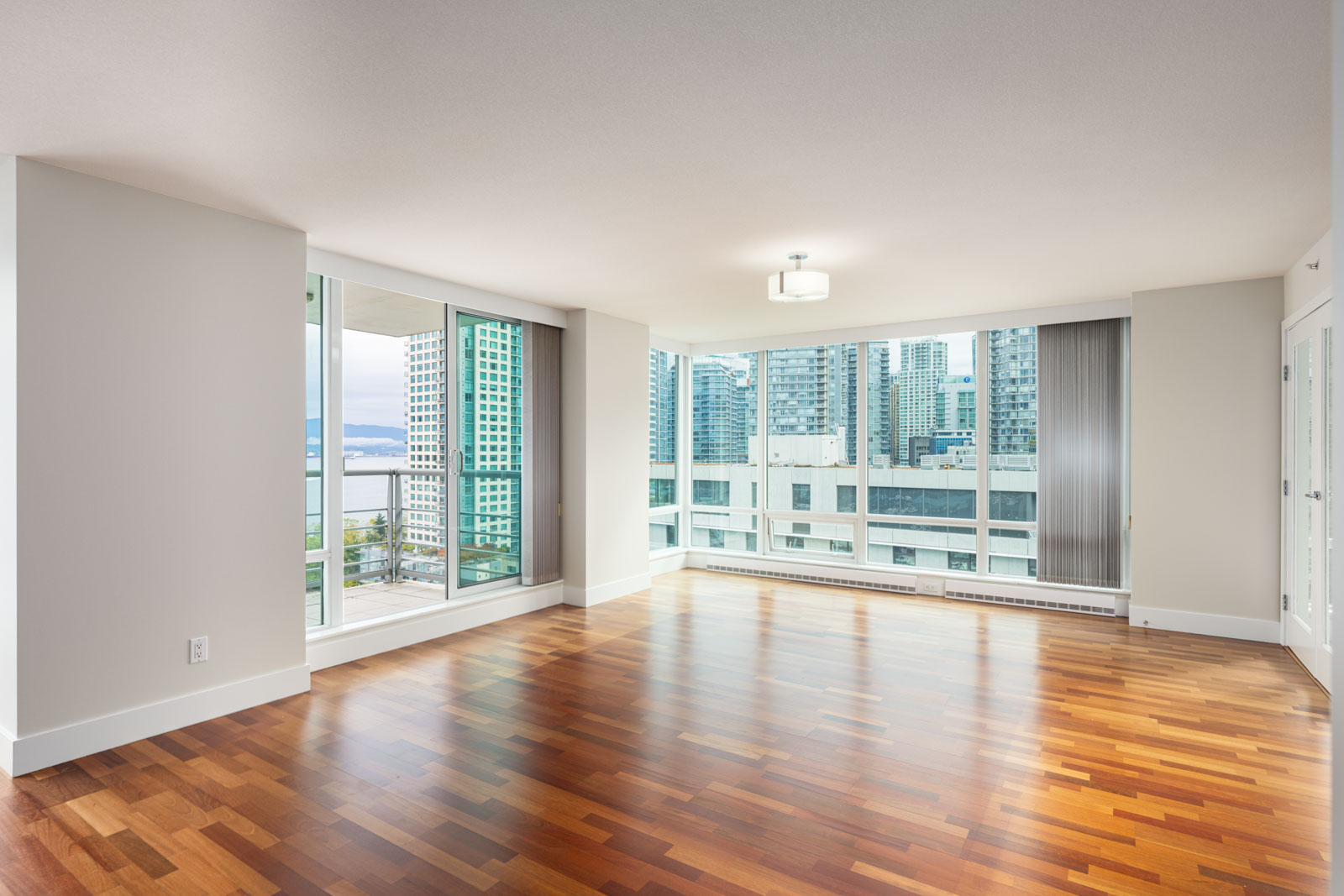 Empty modern apartment living room with hardwood floors, large windows, a balcony, and a view of city buildings and water.