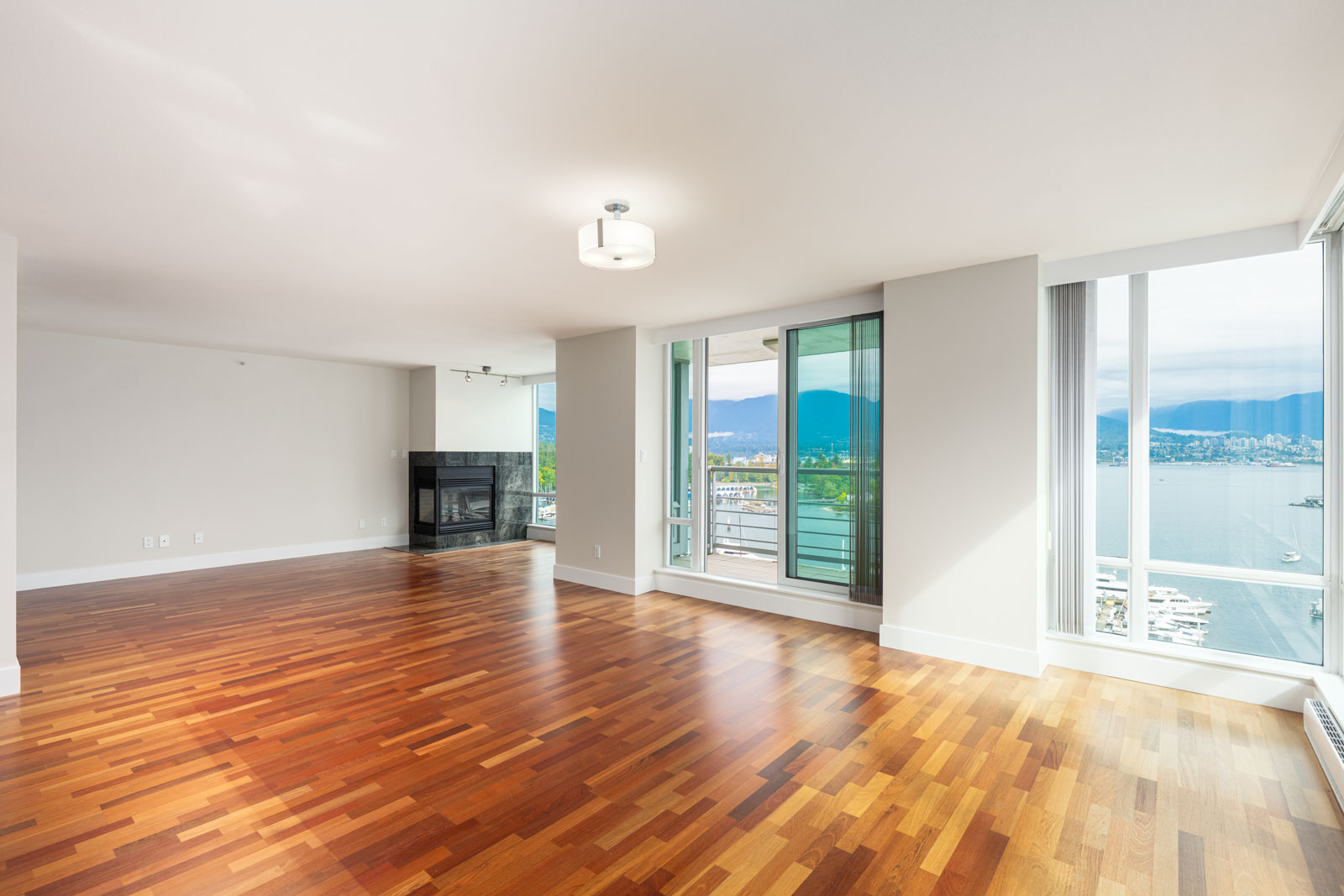 Spacious empty living room with hardwood floors, large windows, sliding glass door to a balcony, and a view of water and mountains.