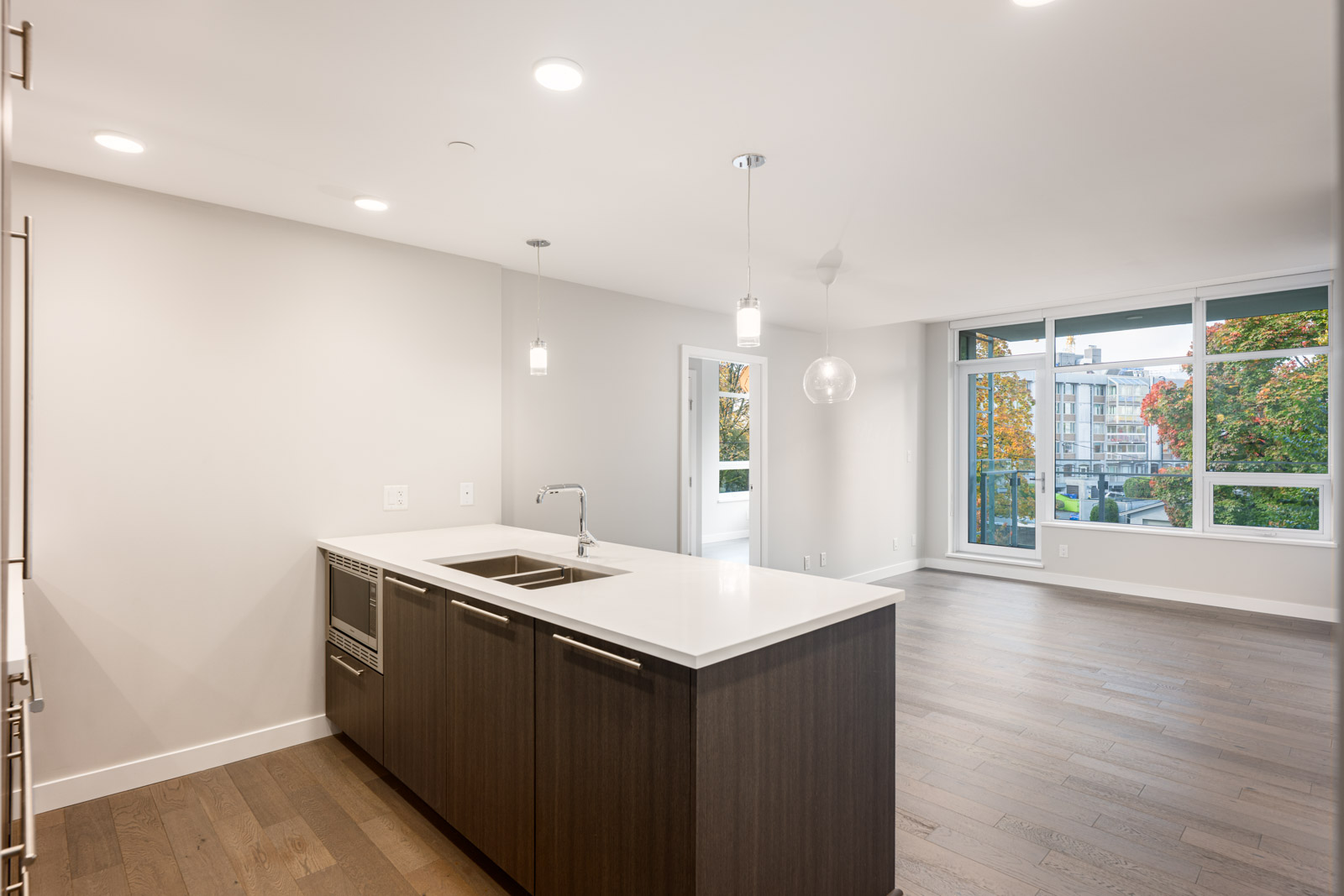 Modern, unfurnished open-plan kitchen and living area with wood flooring, large island, pendant lights, and floor-to-ceiling windows overlooking a cityscape with trees.