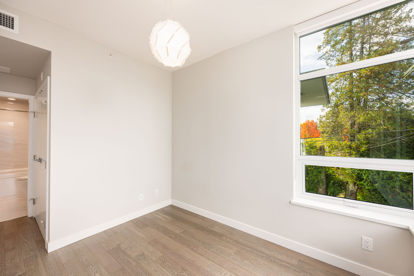 Empty room with light-colored walls, hardwood floor, modern ceiling light, and a large window showing green and autumn trees outside.