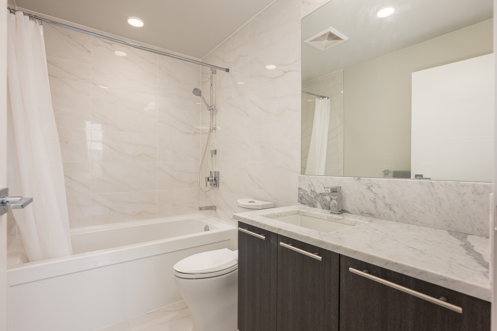 Modern bathroom with a bathtub and shower, toilet, large mirror, marble countertop, and dark wood cabinets. The walls and floor feature light-colored tiles.