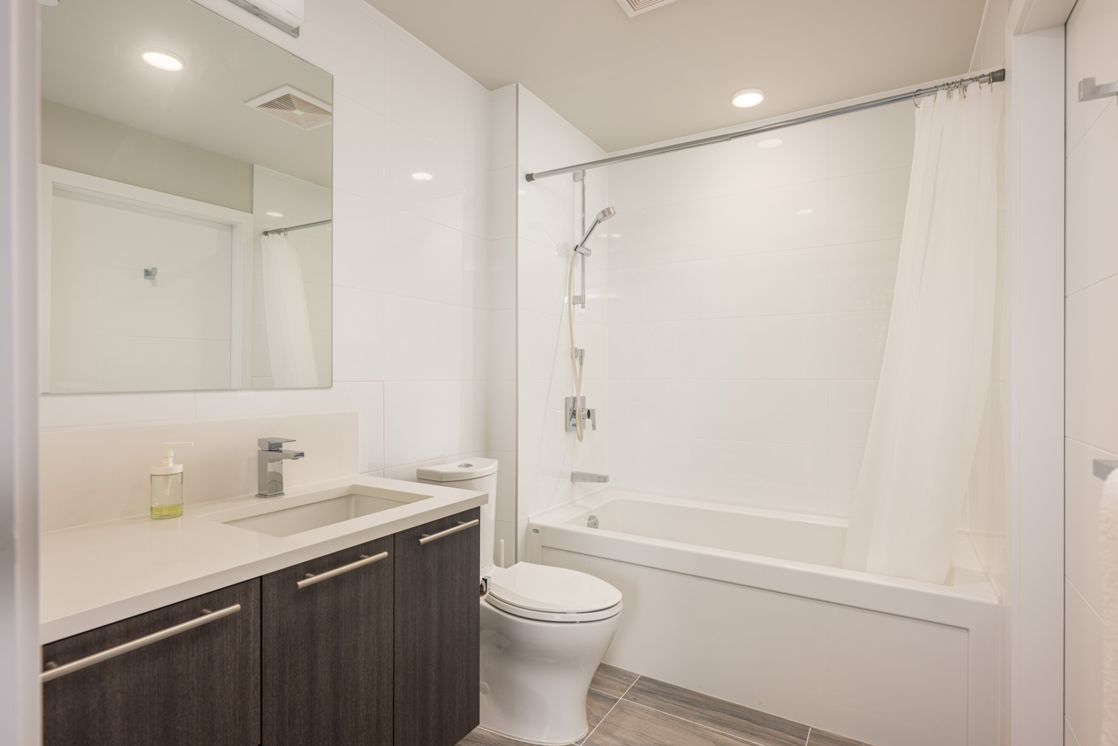 Modern bathroom with a large mirror, dark wood vanity, toilet, and a bathtub with a shower and white curtain. The room features white walls and light wood-look tile flooring.
