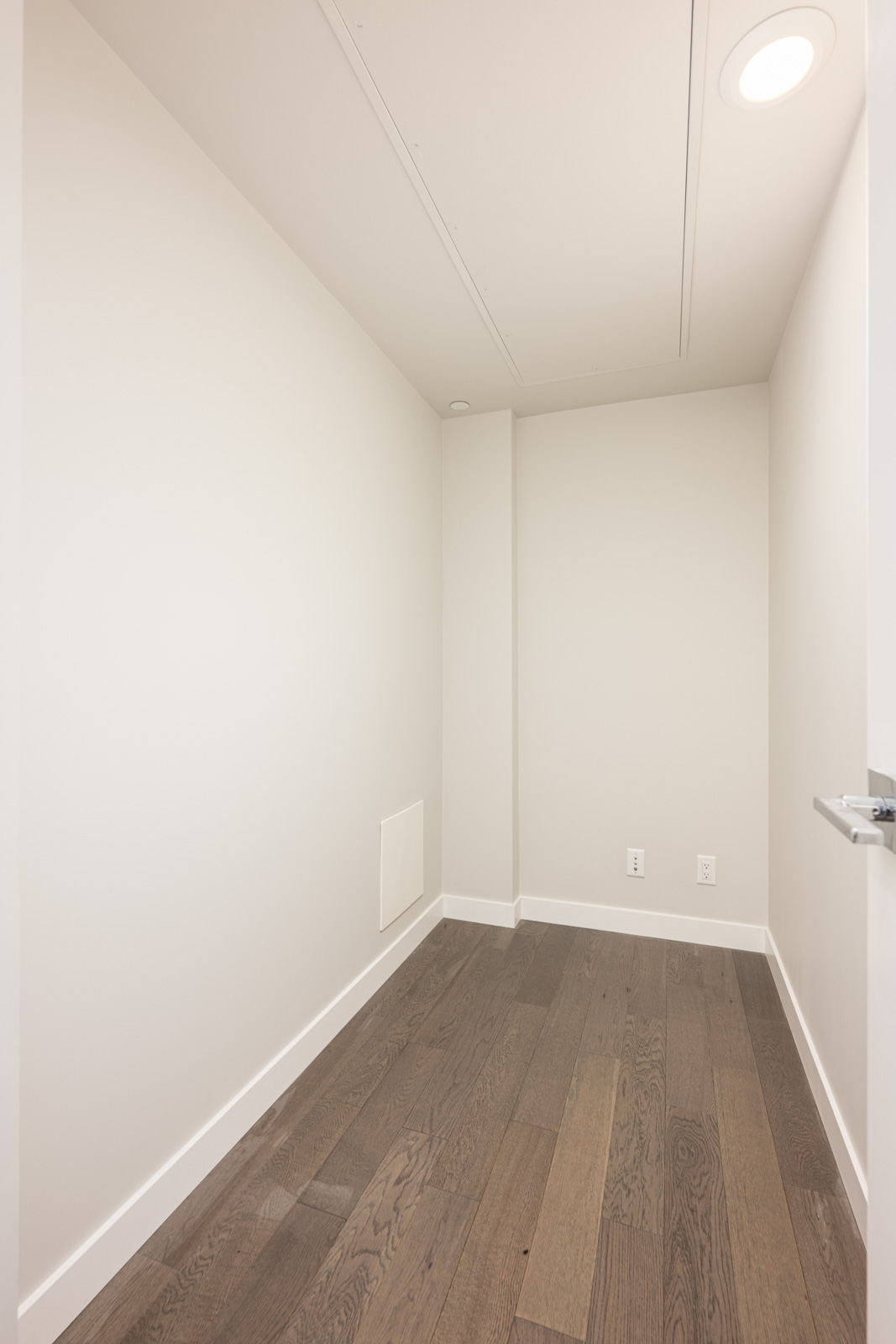 A small, empty room with white walls, wood flooring, and a ceiling light; electrical outlets are visible on the far wall.