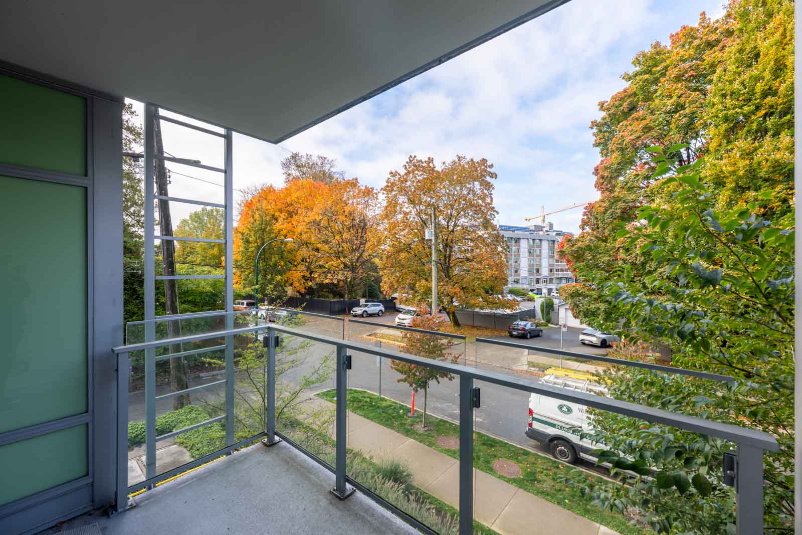 View from a modern balcony overlooking a street lined with trees showing autumn foliage, parked cars, and a construction crane in the distance.
