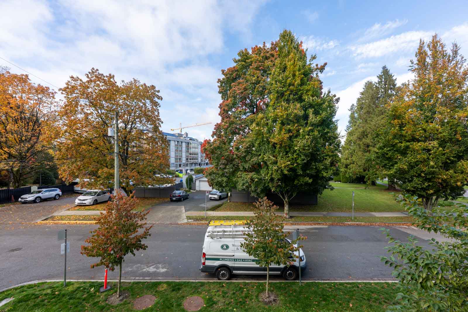 A white service van is parked on a street lined with trees, with cars in a nearby parking lot and buildings visible in the background under a partly cloudy sky.