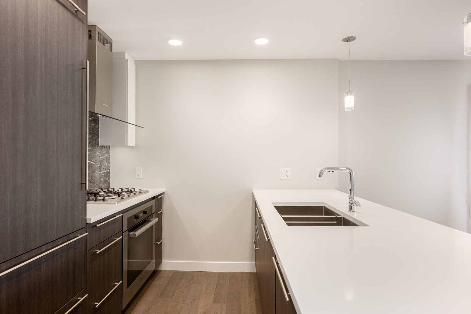 Modern kitchen with dark wood cabinets, stainless steel appliances, white countertops, gas stove, built-in oven, and a pendant light above a sink on an island.