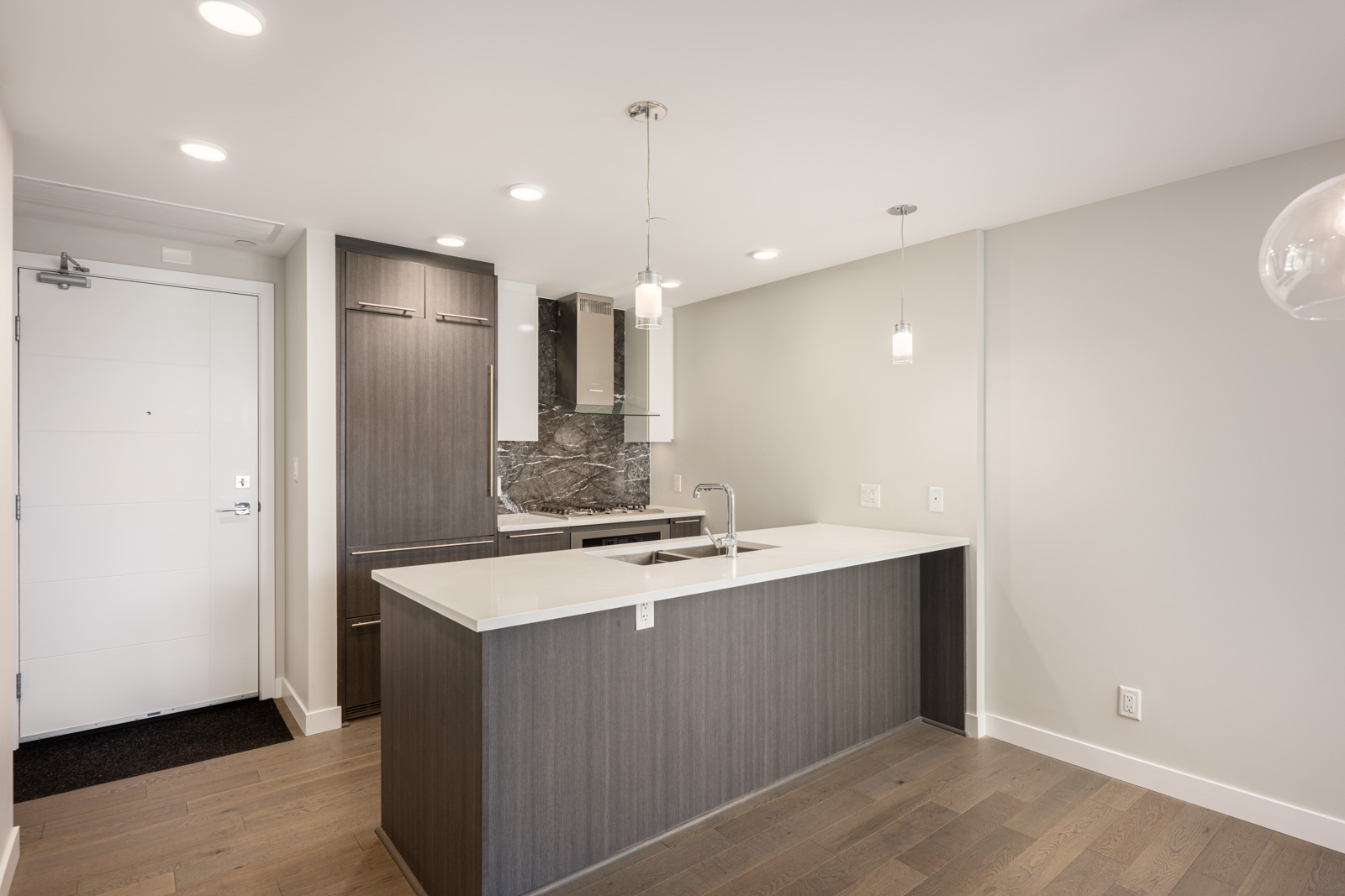 Modern kitchen with dark wood cabinets, a marble backsplash, built-in appliances, and a white island countertop under pendant lighting. White walls and wood flooring complete the space.