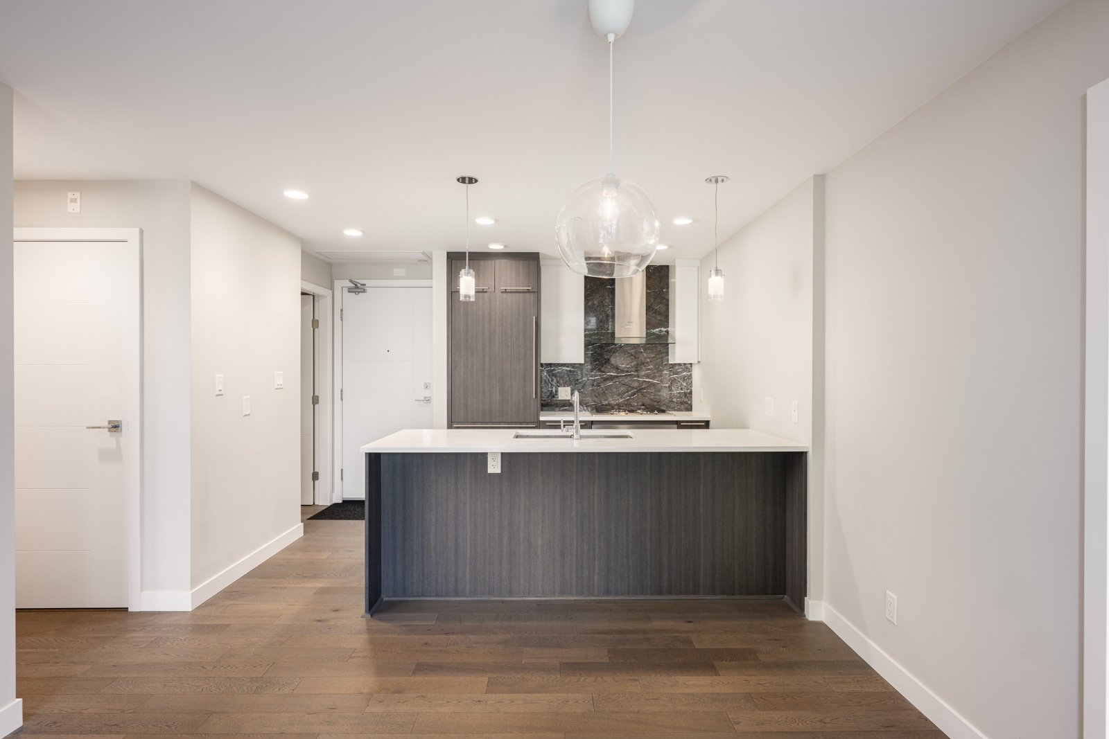 Modern kitchen with a white island, pendant lights, dark wood cabinetry, built-in appliances, marble backsplash, and hardwood floors in a bright, minimalistic interior.